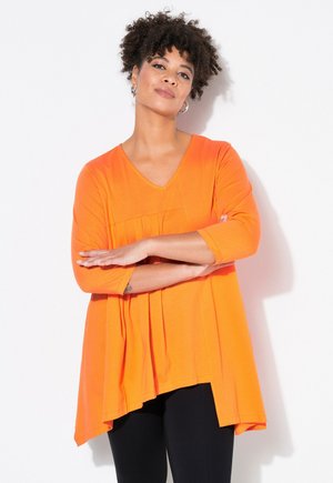 Orange V-hals top med trekvartærmer, A-line silhuet, plisseringer foran og en asymmetrisk kant, parret med sorte bukser.
