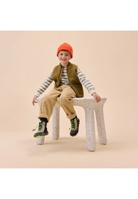 Bunter Skulpturenhocker mit abgerundeten Beinen, gefertigt aus fleckigem Material, unterstützt ein Kind in einer olivfarbenen Weste, gestreiftem Shirt und orangener Mütze.