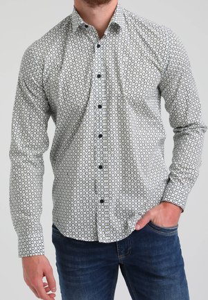 Homme portant une chemise blanche boutonnée avec un motif circulaire noir et un jean bleu foncé, une main dans la poche, sur un fond uni.