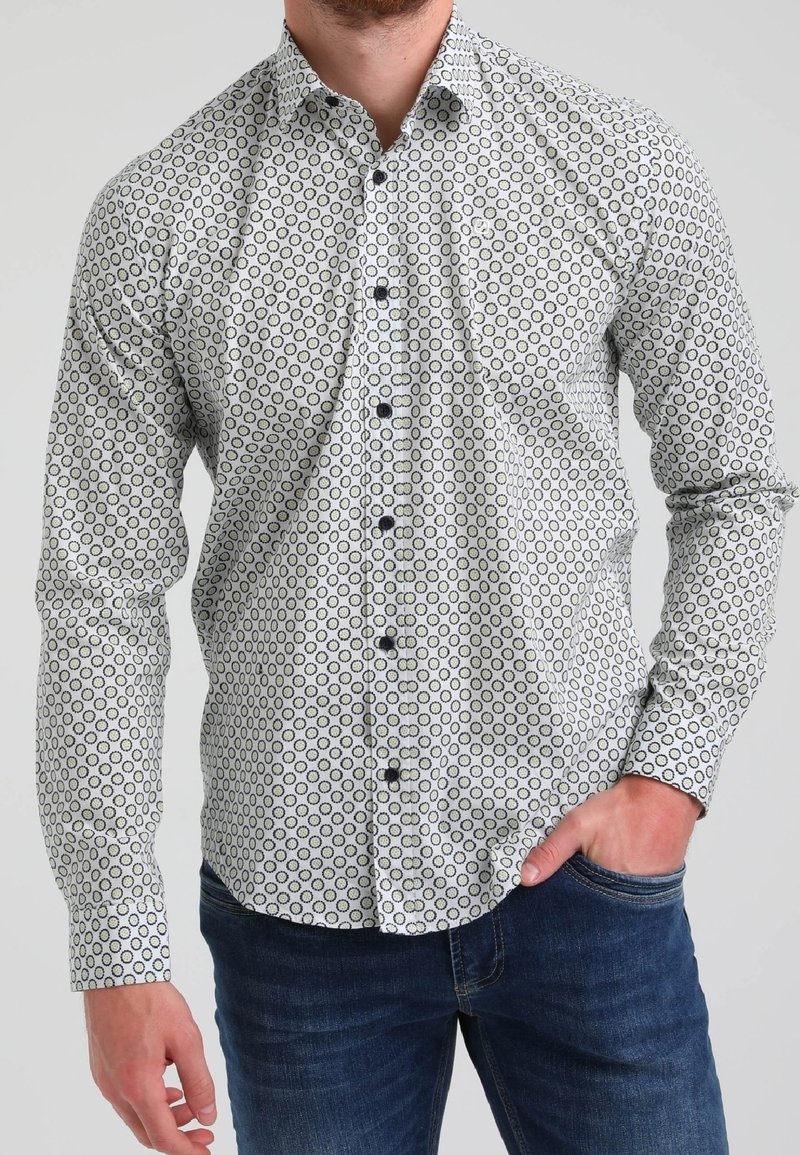 Homme portant une chemise blanche boutonnée avec un motif circulaire noir et un jean bleu foncé, une main dans la poche, sur un fond uni.