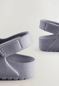 Harmaat kumisaappaat, joissa on teksturoitu päällinen, säädettävä hihna ja pehmeä pohja. Muotoilussa on korotettu kuvio pohjassa.