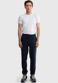 Muž stojící, oblečený v jednobarevném bílém tričku, tmavě modrých teplákových kalhotách s šňůrkou a tmavě modrých, bílých a šedých teniskách na světlém pozadí.