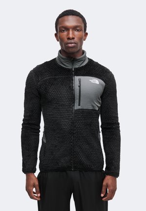 Homme portant une veste zippée noire texturée avec une poche poitrine grise et le logo The North Face, debout devant un fond clair uni.