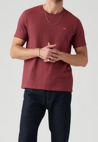 Maroon cotton t-shirt with short sleeves, round neckline, and small logo detail on the chest. Worn with dark blue denim jeans.
