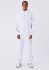 Man staat rechtop en draagt een witte hoodie en bijpassende sweatpants met lichtblauwe piping en witte sneakers, tegen een effen lichte achtergrond.