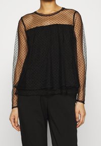 Svart transparent topp med prickar, långärmad, rund halsringning, lagerdesign och textilstruktur. För detaljer finns en mjuk, flödande drapering.