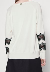 Cremfarbener Pullover mit langen Ärmeln und schwarzen Spitzenakzenten an den Bündchen. Rippstricksaum und Rundhalsausschnitt mit kleinem Rundlogo auf der Rückseite.