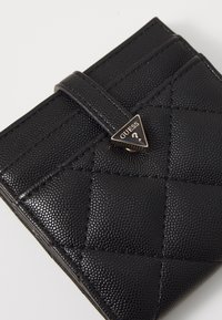 Portafoglio trapuntato nero realizzato in materiale texturizzato, caratterizzato da un accento triangolare con logo GUESS in tono oro e una chiusura a strap.
