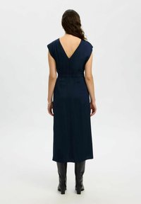 Femme aux longs cheveux noirs portant une robe midi sans manches bleu marine avec un dos en V et des bottes noires jusqu'aux genoux, se tenant devant un fond uni.