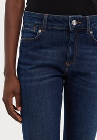 Tmavě modré džíny z denimu s klasickým designem pěti kapes, jemným prošíváním a stříbrným knoflíkem na zapínání v pase.