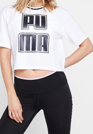 Kobieta ubrana w biały krótki T-shirt marki Puma i czarne legginsy z wysokim stanem, jedną ręką opiera się na biodrze, a drugą trzyma swobodnie wzdłuż ciała.