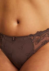 Bruine kantenen slip met bloemmotief, voorzien van een satijnen strikdetail aan de voorkant en een combinatie van doorzichtige en ondoorzichtige materialen.