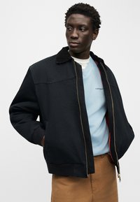 Νεαρός άνδρας με τα χέρια στις τσέπες, φοράει μαύρο μπουφάν με φερμουάρ πάνω από γαλάζιο φούτερ και καφέ παντελόνι, κ οιτάζει προς τα πλάγια.