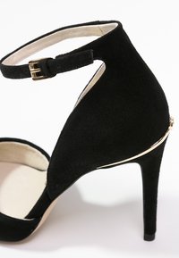 Chaussure à talon haut en suède noir avec bride à la cheville et boucle dorée, ornée d'un fin liseré doré au talon arrière.