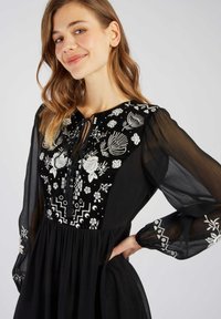 Zwarte jurk met verfijnde witte bloemenborduurwerk op het lijfje, met lange doorzichtige mouwen en een geplooide taille.