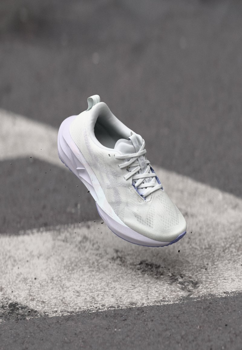 Chaussure de sport blanche avec une tige texturée, un design à lacets et une semelle rembourrée. Les accents comprennent une semelle extérieure violet clair et une languette à l'arrière.