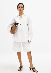 Oversized white button-up shirt with long sleeves paired with white shorts featuring contrasting cuffs. Brown handbag and dark heeled sandals.