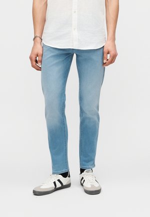 Άνδρας που φοράει ανοιχτό μπλε τζιν στενής γραμμής, λευκό πουκάμισο με κουμπιά, μαύρες κάλτσες και λευκά αθλητικά παπούτσια με μαύρες ρίγες, στέκεται μπροστά σε λευκό φόντο.