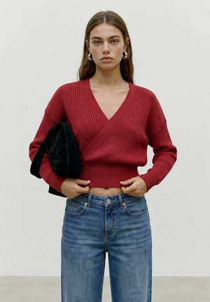 Giovane donna con capelli lunghi indossa un maglione rosso a coste con scollo a portafoglio, jeans blu, orecchini a cerchio d'argento e tiene una borsa nera sotto il braccio.