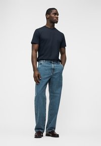 Man standing with hands in pockets, wearing a black t-shirt, blue jeans, and dark brown shoes, against a plain light background.