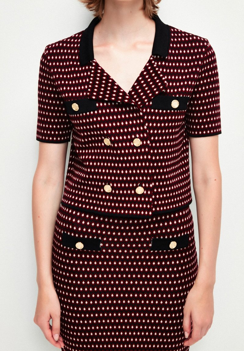 Femme portant une veste à manches courtes rouge et noire à motif et une jupe assortie avec des boutons dorés et une bordure noire.