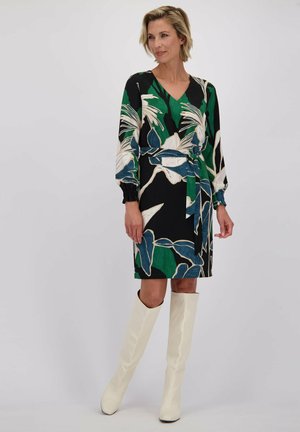 Robe noire avec un motif floral vert et blanc, décolleté en V, manches longues avec des poignets smockés, taille cintrée avec une ceinture assortie, portée avec des bottes blanches jusqu'au genou.