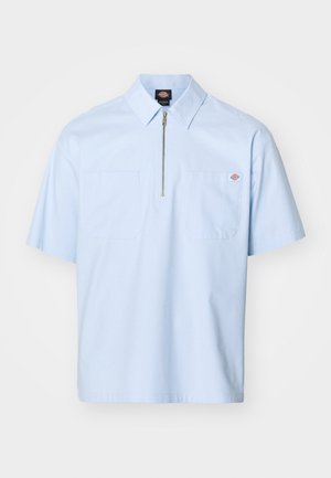 Rövid ujjú világoskék ing, hegyes gallérral, elől cipzárral és két nagy zsebbújtató zsebbel. Simább anyagból készült, laza fazonban.