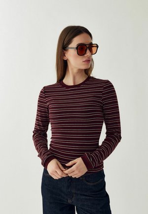 Žena s dlhovlasými hnedými vlasmi, ktorá nosí veľké oranžovo sfarbené slnečné okuliare, bordovú pruhovanú blúzku s dlhým rukávom a tmavomodré rifle, ruky zložené.