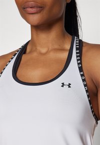 Canottiera atletica bianca con bordi e spalline nere. Presenta la scritta "Under Armour" stampata sulle spalline e un piccolo logo sul petto. Texture liscia.