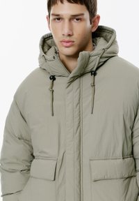 Svetlo olivovo zelená puffa bunda s kapucňou, dvoma prednými vreckami, elastickými sťahovacími šnúrkami a kovovými detailmi. Hladká, textúrovaná tkanina.