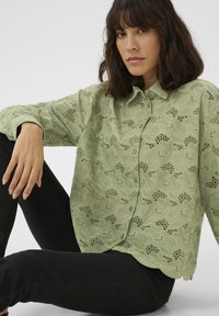 Chemise vert clair avec des motifs floraux en découpes complexes, fermeture à boutons sur le devant et un ourlet légèrement courbé. Assortie à un pantalon noir.