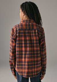 Flannelskjorte i orange, marineblå og hvid tern. Har en knaplukning på kraven, lange ærmer og buet kant. Blød tekstur, afslappet pasform.