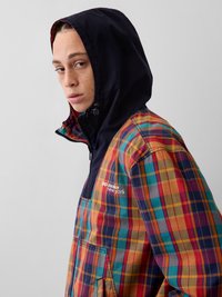 Άτομο με πολύχρωμο καρό μπλεζάκι με κουκούλα και κείμενο «gap awake new york» στο στήθος, στραμμένο ελαφρώς στο πλάι σε απλό φόντο.