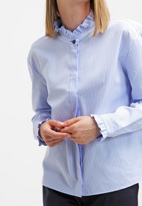 Jasnoniebieska koszula w paski z falbaniastym kołnierzykiem i mankietami, z zapięciem na guziki z przodu oraz gładką teksturą materiału.