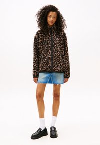 Vrouw met krullend haar, gekleed in een bruine panterprint zip-up jack, denim minirok, witte sokken en zwarte instappers, staand tegen een witte achtergrond.