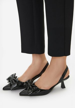Czarne czółenka slingback z lakierowanej skóry, z zakończeniami w szpic i dużymi ozdobnymi kokardami, na stopach w czarnych krótkich spodniach.