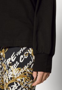 Top de algodón negro de manga larga con puños elásticos, combinado con leggings estampados que presentan texto dorado y blanco sobre un fondo oscuro.