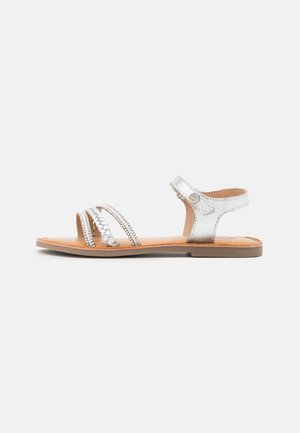 Sølvmetallic sandal har tre stropper med dekorative detaljer, en flad brun sål og en ankelrem med spænde.