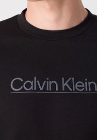 T-shirt in cotone nero con scollo rotondo, caratterizzato da un logo ricamato "Calvin Klein" grigio a contrasto sul petto.