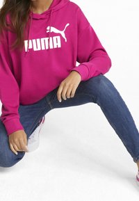 Sudadera magenta con un logo blanco de Puma, combinada con jeans azul oscuro. La tela parece suave y el diseño incluye un bolsillo frontal y una capucha ajustable.