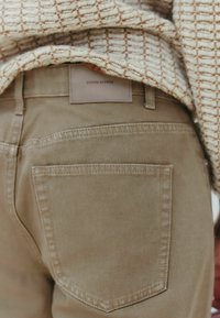 Beežid puuvillased püksid tagatasku ja nahklabeliga, millel on kiri "Denim Supply". Kangas on tekstuuriga pinna ja sirge lõikega.