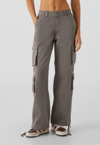 Pantalon cargo gris avec un design à jambes droites, plusieurs poches sur les côtés et une texture légèrement froissée. Porté avec des baskets décontractées.