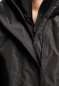 Zwarte ritsjack met een textuurstof, twee zijritsen en een hoge kraag. Het materiaal heeft een subtiel ruitpatroon.