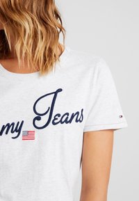 Šedé tričko s kulatým výstřihem, které má námořnickou vyšívanou náplast "my Jeans" a malou grafiku americké vlajky. Zahrnuje logo na rukávu.