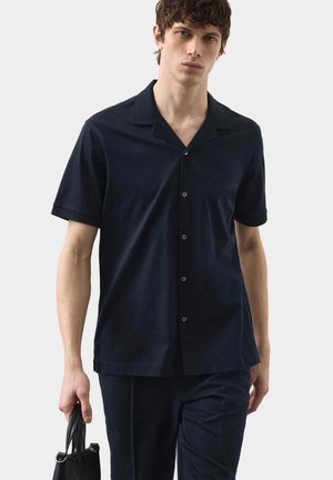 Joven con camisa de manga corta azul marino abotonada y pantalones sostiene una bolsa de cuero negra, de pie frente a un fondo liso y claro.