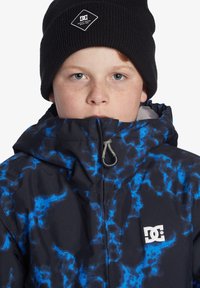 Svart jacka med blå blixtmönster, hög krage och blixtlås framtill. Har DC-logotyp på bröstet och en texturerad svart stickad mössa.