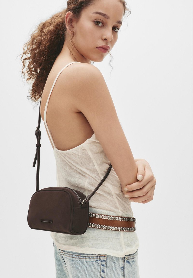 Jeune femme aux cheveux bouclés portant un débardeur blanc transparent, une ceinture cloutée, un jean bleu clair et un petit sac bandoulière noir.
