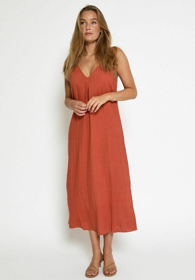 Robe maxi couleur rouille avec un décolleté en V, des bretelles spaghetti et une coupe ample. Fabriquée en tissu texturé, atteignant le milieu du mollet. Sandales visibles en dessous.