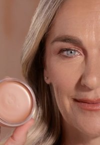 Jasnołososiowa krem w przezroczystym, okrągłym pojemniku trzymanym przez kobietę. Gładka tekstura, refleksyjna powierzchnia, naturalny makijaż, neutralne tło.