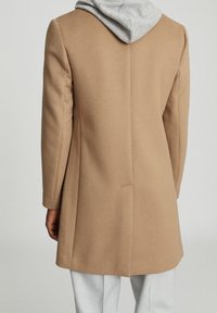 Man in een camel-kleurige overjas over een lichtgrijze hoodie, gecombineerd met lichtgrijze broeken, van achteren gezien tegen een effen achtergrond.